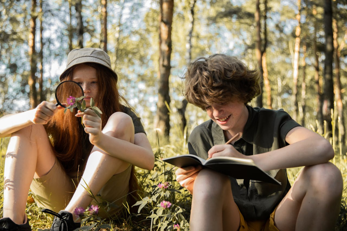 Girl and boy in summer camp gear, in woods, looking at plants and writing in journal