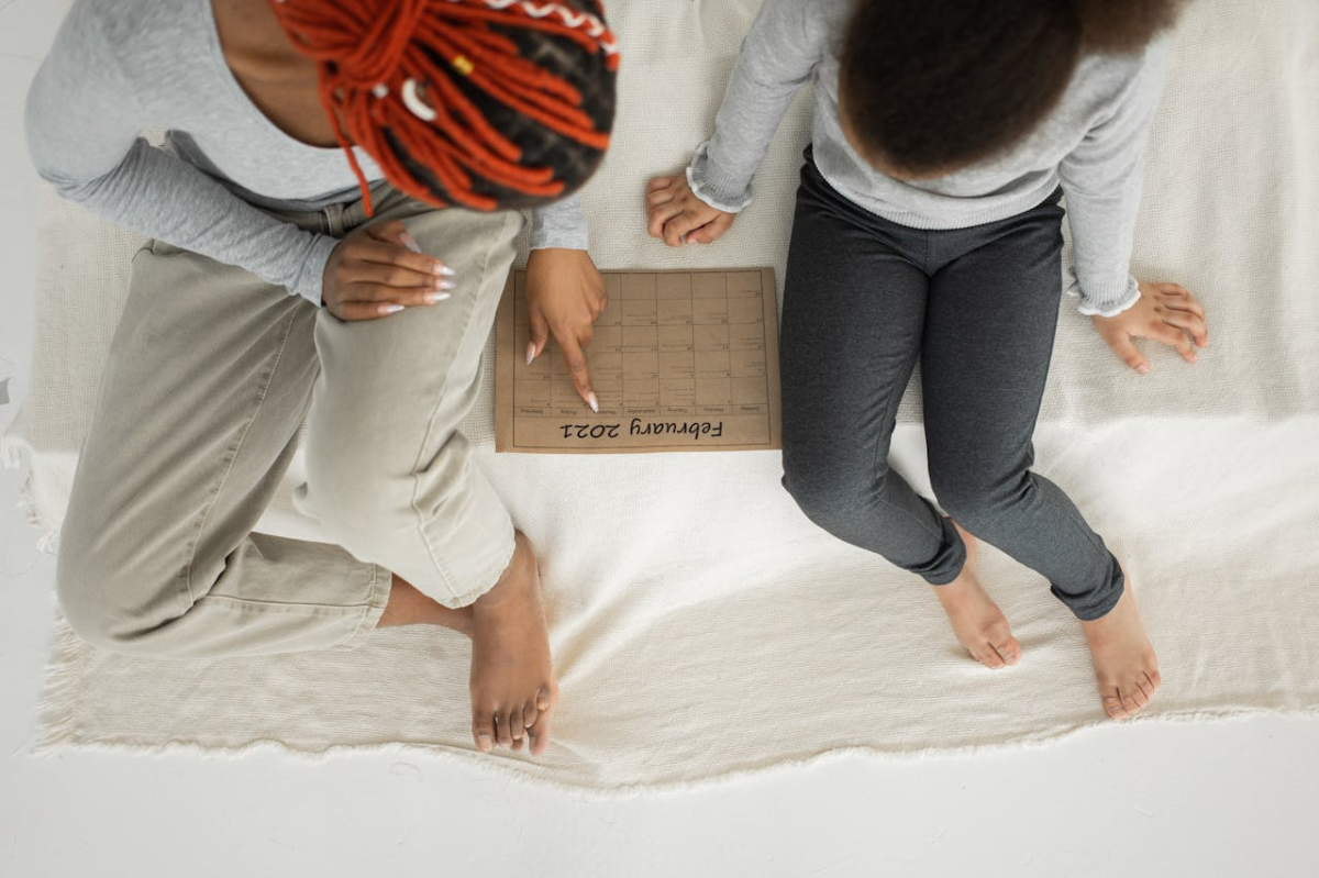 Black mother and daughter sitting on bed pointing at a calendar of events